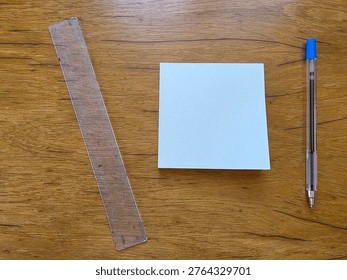 A top-down, minimalist composition features three common office essentials arranged on a warm, textured wooden surface. In the center sits a blank, square white notepad, ready for notes or sketches. To its left, a transparent plastic ruler lies at a slight angle, showing signs of use with faint markings. To the right, a classic clear ballpoint pen with a blue cap and tip is placed vertically. The natural grain of the wood provides a rustic background, creating an atmosphere of organized productivity, brainstorming, or academic study.
