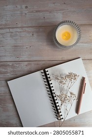 Top-Down Open Notebook And Pink Pen With Cream Lit Candle And Gypsophila On Neutral Coloured Wooden Table
