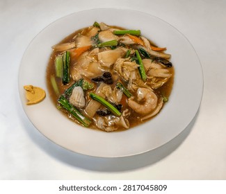 A top-down, overhead shot capturing a plate of Japanese-style Gomoku Ankake Yakisoba (五目あんかけ焼きそば), featuring crispy pan-fried noodles smothered in a thick, savory starchy sauce with mixed seafood and vegetables. The dish includes shrimp, squid, pork, wood ear mushrooms, napa cabbage, carrots, and leafy green vegetables, served in a round white shallow bowl with a dollop of yellow karashi mustard on the rim. Set against a clean white table surface, this image is ideal for stock media covering Japanese-Chinese cuisine (Chuka), noodle dishes, lunch menus, and Asian culinary photography.