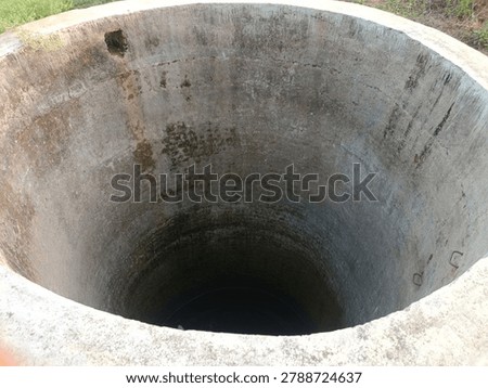 Top-down perspective of a deep abandoned concrete well showing extreme water scarcity. The circular well walls have moss, algae stains, and weathering marks from years of use. Only a small dark patch of water visible at the very bottom, showing severe groundwater depletion. Rusty iron foot-holds visible on side wall.
