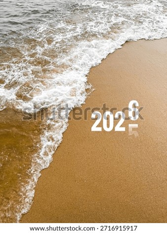 A top-down perspective of ocean waves with white sea foam washing onto a sandy beach. The image features a creative typography overlay transitioning from ‘2025’ to ‘2026’, symbolizing the New Year. This high-angle landscape combines natural elements with seasonal messaging, ideal for travel, resolution-themed, or conceptual editorial content.