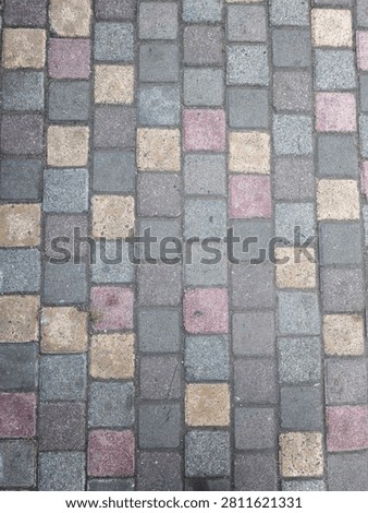 top-down photograph shows a section of a cobblestone or paver walkway. The path is made of small, square paving stones arranged in neat vertical columns.
The stones feature a textured, slightly weathered finish with a muted color palette. While the majority of the pavers are varying shades of cool gray and charcoal, they are interspersed with randomly placed accent stones in faded shades of dusty pink, burgundy, and warm beige-gold. Thin gaps filled with dark dirt or sand separate the individual blocks, creating a distinct grid pattern.