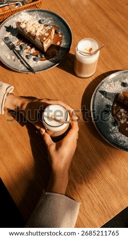 A top-down shot captures a person's hands holding a coffee drink, alongside plates of cake and coffee on a wooden table, bathed in sunlight.