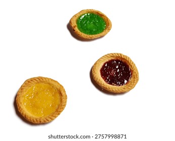 A top-down, slightly elevated perspective photograph featuring three small Indonesian milk tarts (Pai Susu) arranged in a triangular pattern against a clean, brilliant white background. The tart at the top features a vibrant green jam topping, the center-left tart has a bright yellow jam, and the bottom-right tart displays a deep red strawberry jam. All tarts possess a golden-brown, delicately crimped, buttery crust, highlighting the glossy, glistening fruit fillings. This image is perfect for culinary blogs, dessert menus, bakery advertisements, and articles focused on Asian sweets and souvenirs.