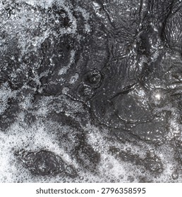 A top-down, square format shot of dark, murky water churning heavily with muddy currents, oily ripples, and patches of thick white foam and bubbles.