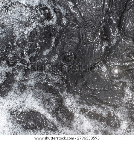A top-down, square format shot of dark, murky water churning heavily with muddy currents, oily ripples, and patches of thick white foam and bubbles.