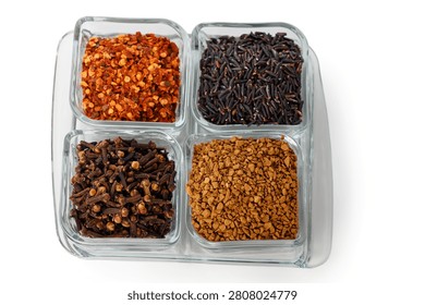 A top-down studio shot of four square glass bowls arranged together, containing red chili flakes, black forbidden rice, whole cloves, and instant coffee granules, isolated on a white background.