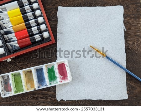 Top-down view of an art setup featuring watercolor paints, paint tubes, a paintbrush, and a blank handmade textured paper sheet on a dark wooden table.