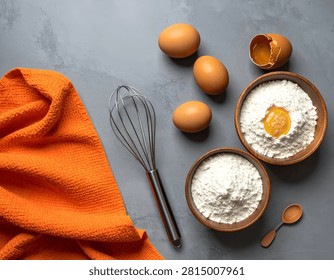 A top-down view of baking ingredients including eggs and flour in bowls, a whisk, and an orange cloth on a grey textured background.