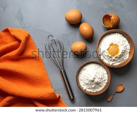 A top-down view of baking ingredients including eggs and flour in bowls, a whisk, and an orange cloth on a grey textured background.