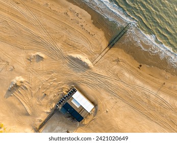 Top-down view of a beach with waves breaking on the sandy shoreline and a beach hut a short distance from the sea