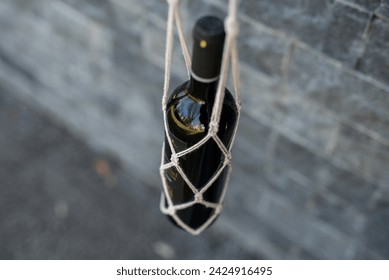 Top-down view: Black wine bottle with red wax-sealed cork in macramé holder, hanging beside a grey brick wall. A stylish composition blending elegance and rustic charm. - Powered by Shutterstock