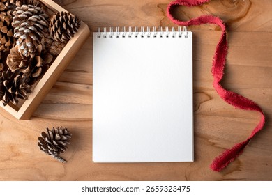 Top-down view of a blank spiral notepad on a wooden surface with pinecones and red ribbon, perfect for winter or holiday themes, writing lists, or seasonal messages.