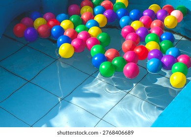 Top-down view of brightly colored plastic play balls floating in a sunlit tiled swimming pool. High contrast lighting casts deep shadows under the clear water, emphasizing a playful, graphic summer aesthetic.