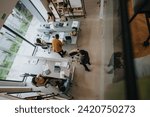 Top-down view of a bustling open-plan office with diverse professionals collaborating at desks and in casual seating areas, exemplifying teamwork in a modern workplace setting.