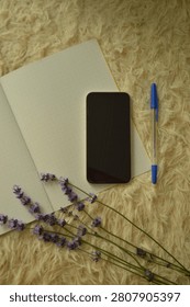 A top-down view captures a modern smartphone, open dotted notebook, blue pen, and fresh lavender sprigs arranged on a soft, textured surface