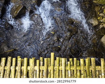 Top-down view of a clear mountain stream flowing over natural rocks beneath a rustic bamboo bridge. Fresh running water, natural textures, and tranquil forest scenery create a peaceful outdoor landscape.