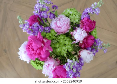 A top-down view of a colorful bouquet featuring pink peonies, purple flowers, and green hydrangeas arranged on a wooden surface, showcasing a vibrant floral design. - Powered by Shutterstock