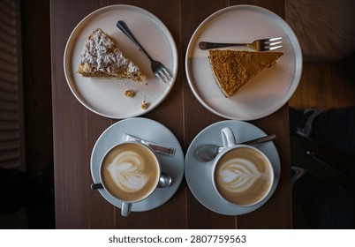 Top-down view of a cozy cafe date featuring two cups of fresh cappuccino with latte art and two delicious slices of cake on white plates over a dark wooden table.