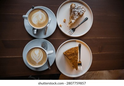 Top-down view of a cozy cafe date featuring two cups of fresh cappuccino with latte art and two delicious slices of cake on white plates over a dark wooden table.