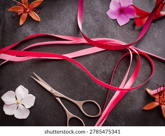 A top-down view of a dark textured surface decorated with satin pink and red ribbons, colorful loose flowers, and a pair of vintage craft scissors.