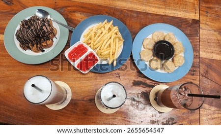 A top-down view of a delicious spread of snacks and drinks on a rustic wooden table, featuring waffles with chocolate ice cream, french fries with ketchup, fried snacks, and refreshing iced beverages.