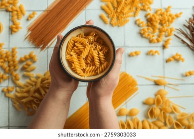 A top-down view of different uncooked pasta types scattered on a tiled surface with a bowl of fusilli held in hands
