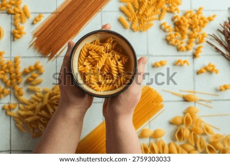 A top-down view of different uncooked pasta types scattered on a tiled surface with a bowl of fusilli held in hands