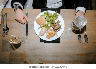 top-down view of a dining table setting. A person in formal attire, marked by a tuxedo, is seated and appears to be enjoying a meal. On the plate, there is a variety of food including meat,