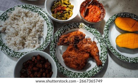 A top-down view of a dining table with a variety of Indonesian dishes, including rice, fried chicken, sauteed vegetables, mango, and a bowl of fried tempeh.