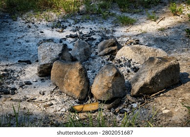 Top-down view of a disused fire pit with rocks, ash, and charcoal on sandy earth, surrounded by grass. A professional nature stock photo.