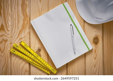 Top-down view of engineering notepad with pen, yellow folding ruler and white safety helmet on wooden surface. Construction, drafting, architectural planning, measurement, structured thinking