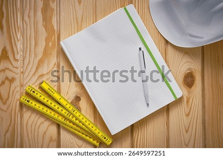 Top-down view of engineering notepad with pen, yellow folding ruler and white safety helmet on wooden surface. Construction, drafting, architectural planning, measurement, structured thinking