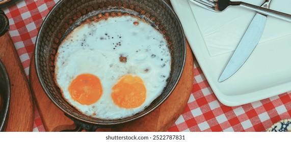 Top-down view of fried eggs in a pan on a red checkered tablecloth, accompanied by a plate and cutlery in daylight