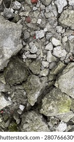 A top-down view of a ground surface covered in a mix of jagged gray rocks, crushed concrete, and small stone fragments. The texture is rough and uneven, featuring various sizes of debris ranging from large, moss-covered boulders to fine gravel and small pebbles. The color palette is dominated by shades of gray, slate, and earthy brown, with subtle hints of green moss and small flecks of red brick embedded within the fractured