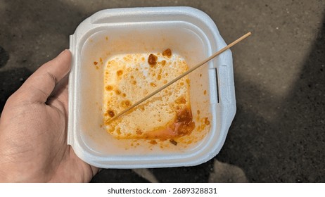 A top-down view of a hand holding an empty and dirty white styrofoam food container with leftover spicy sauce and a wooden skewer. A concept for being finished, food waste, or the aftermath of eating.