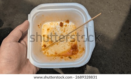 A top-down view of a hand holding an empty and dirty white styrofoam food container with leftover spicy sauce and a wooden skewer. A concept for being finished, food waste, or the aftermath of eating.