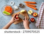 A top-down view of hands grating a red apple onto a wooden cutting board surrounded by ingredients for a salad.