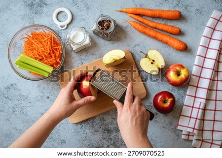 A top-down view of hands grating a red apple onto a wooden cutting board surrounded by ingredients for a salad.