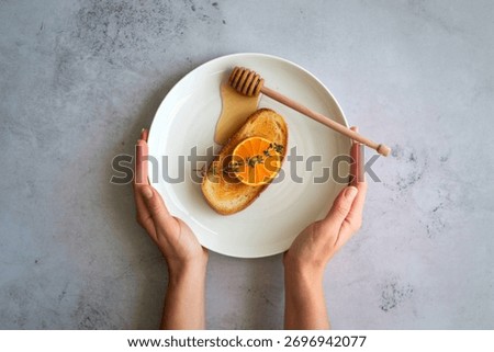 A top-down view of hands presenting a white oval plate topped with toast, fresh orange slices, and a honey dipper drizzling honey.