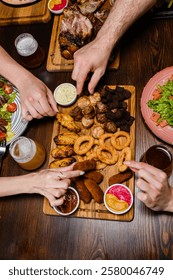 A top-down view of hands reaching for assorted appetizers on a wooden platter, including onion rings, chicken wings, and sausages. Beer and salads are on the table, creating a cozy dining scene.