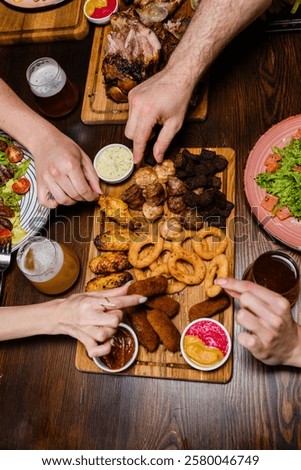A top-down view of hands reaching for assorted appetizers on a wooden platter, including onion rings, chicken wings, and sausages. Beer and salads are on the table, creating a cozy dining scene.