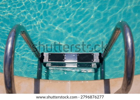Top-down view of hotel pool entry with steel handrails framing the submerged pool ladder step, pool water showing mosaic tile on the bottom, beige stone pool coping visible near the ladder