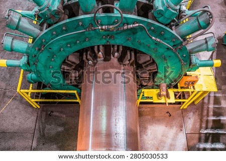 A top-down view of a large industrial machine, featuring a central metal pipe passing through a green circular clamp mechanism equipped with hydraulic actuators, likely used for heavy-duty pipeline manufacturing or repair processes.