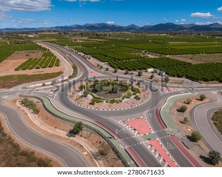 Top-down view of a large modern roundabout with red-marked cycle lanes surrounded by orange groves, agricultural fields and mountain range in Spain