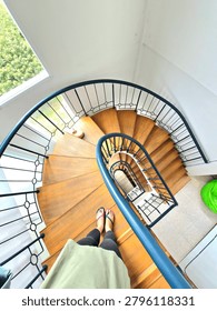 a top-down view looking down a winding spiral staircase made with light-colored wooden steps and a dark-blue painted metal handrail. The lower half of a person wearing black leggings, a green shirt, and blue and pink flip-flops is visible at the very top of the stairs, with their feet planted on the first step. The staircase curves away to the bottom, ending on a light-tiled floor next to a green bean bag chair. Natural light floods in from a tall window at the far left side of the staircase, revealing trees outside. Small, dark, abstract figures are dotted along the metal handrail as it coils down to the bottom
