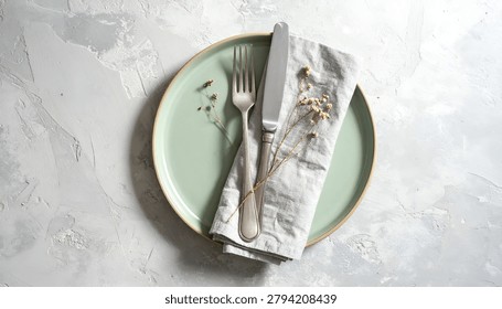 A top-down view of a minimalist table setting featuring a mint green plate, a folded linen napkin, and polished silverware on a light gray textured surface.
