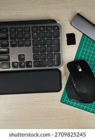 Top-down view of a modern workspace featuring a sleek keyboard, ergonomic mouse, SD card, USB hub, and cutting mat arranged neatly on a light wooden desk.