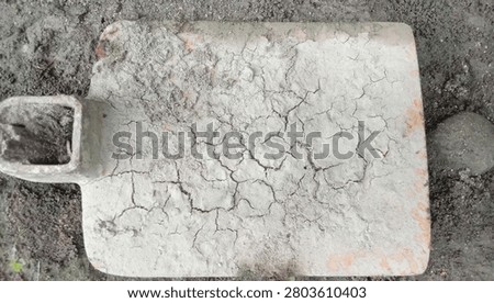 A top-down view of an old metal hoe blade resting on the ground in Its surface is covered with a thick, textured layer of dry, heavily cracked mud and dirt.