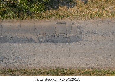 A top-down view of an old worn asphalt road featuring cracks, patched sections and dry grass along the edges, illustrating aging infrastructure and rural road conditions.